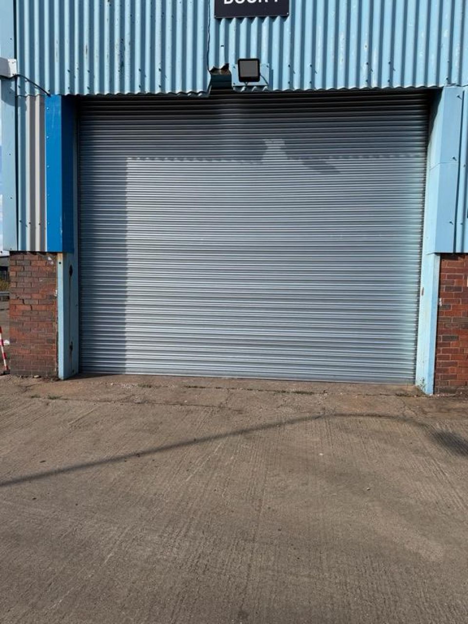  Closed silver roller shutter on the exterior of a building with blue metal cladding and brick walls.